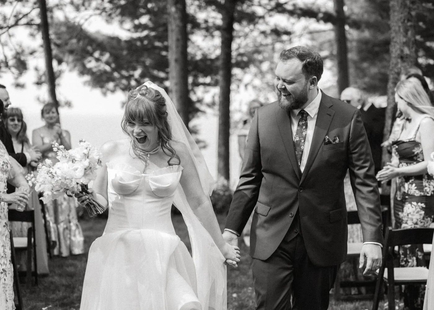 Becca and Justin walking back down the aisle hand in hand on their wedding day, Becca laughing with her bouquet raised, in black and white