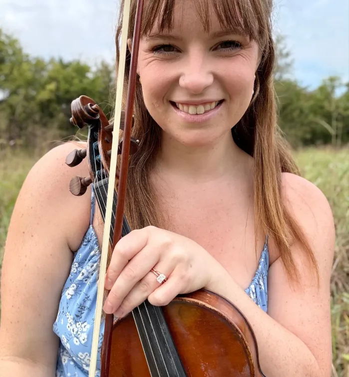 Becca Augustine smiling and holding her violin outdoors