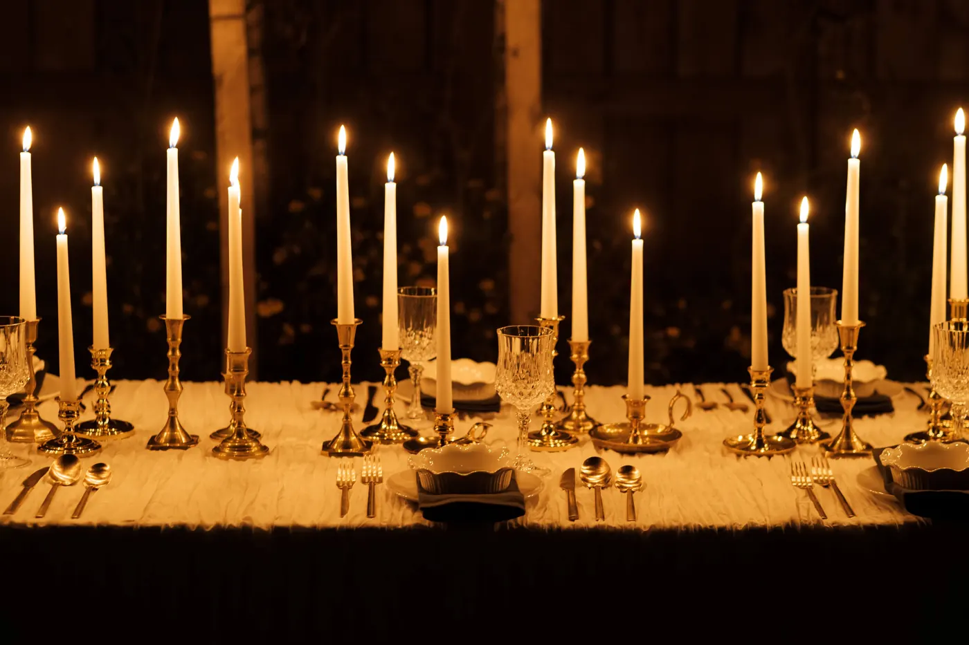 A candlelit table lined with single brass candlesticks of different heights holding white tapers