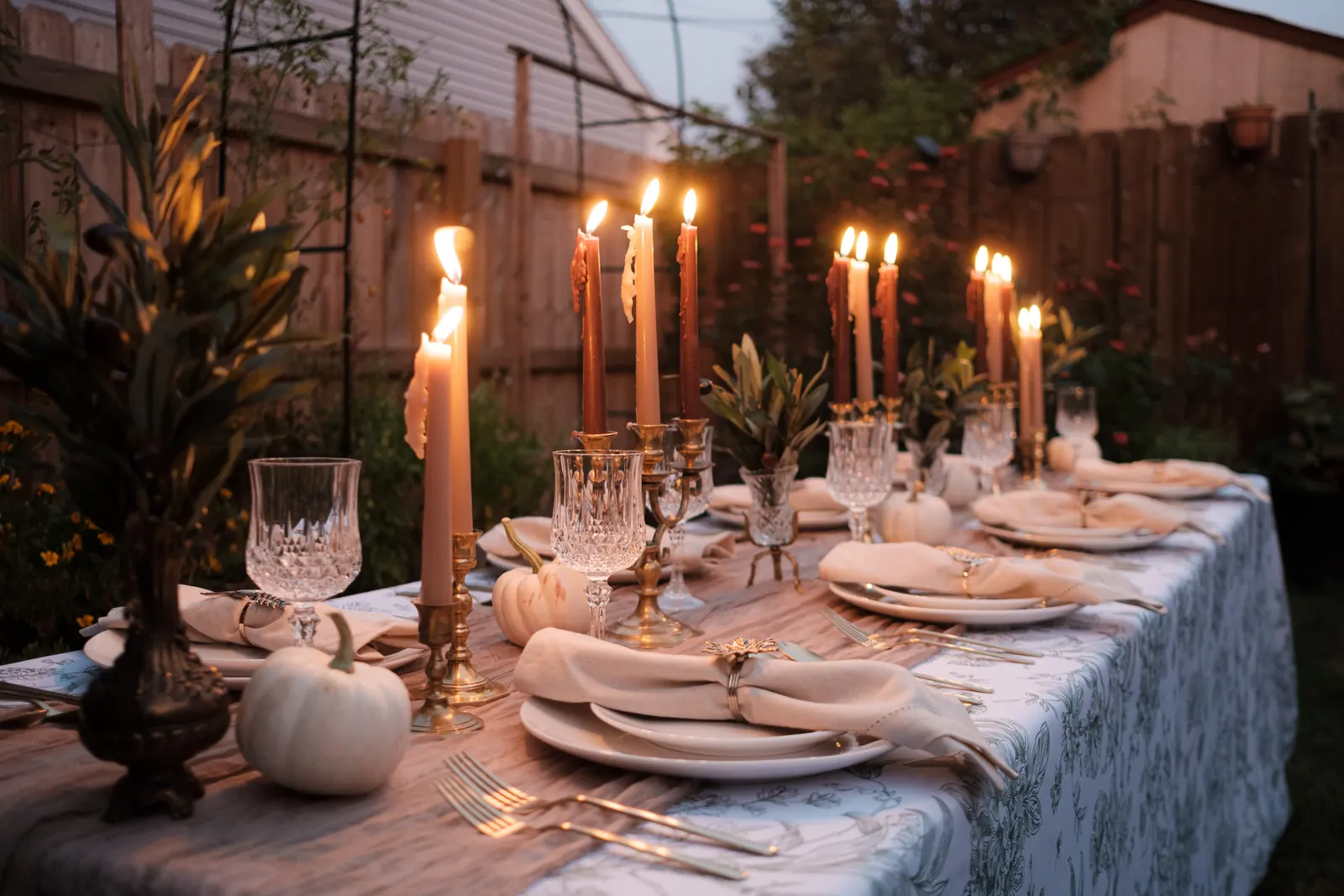 A full harvest table at dusk lined with 3 arm brass candelabras holding burnt orange and cream tapers, with crystal goblets, white pumpkins and toile linen