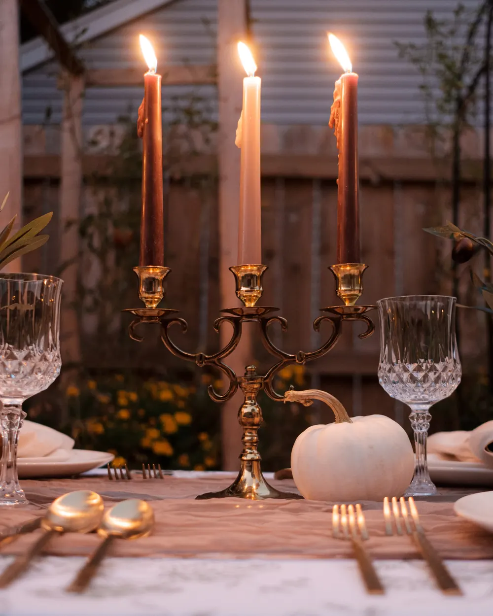 A 3 arm brass candelabra with lit burnt orange and cream tapers between crystal goblets and a white pumpkin
