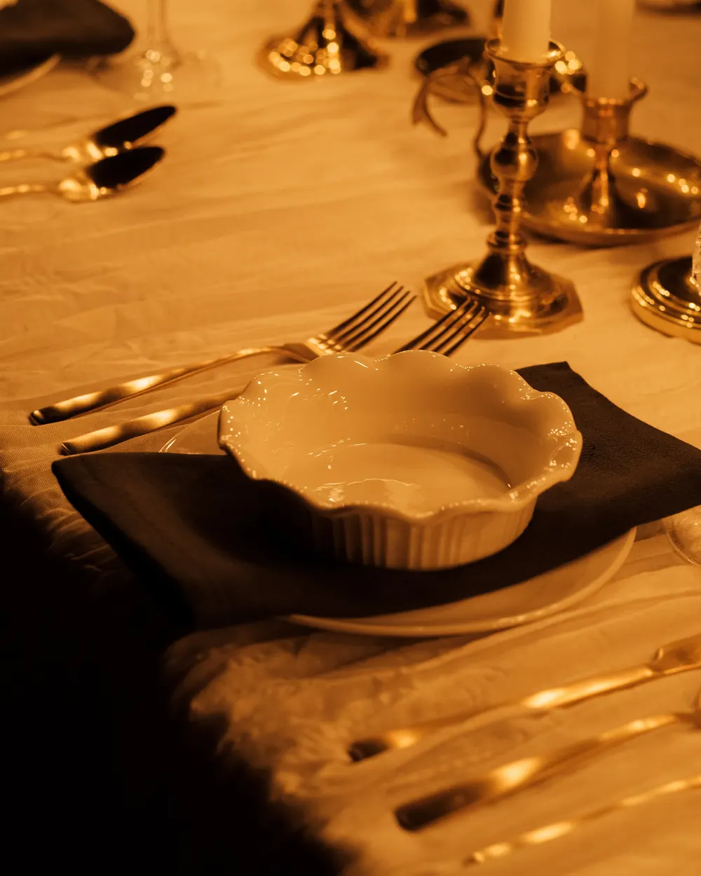 A white ruffled-rim dessert ramekin on a dark napkin, with gold flatware and brass candlesticks