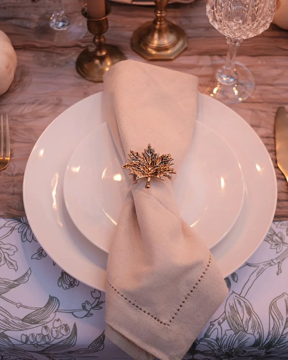 White round dinner plates stacked in a place setting with a cream napkin and gold leaf napkin ring on toile linen