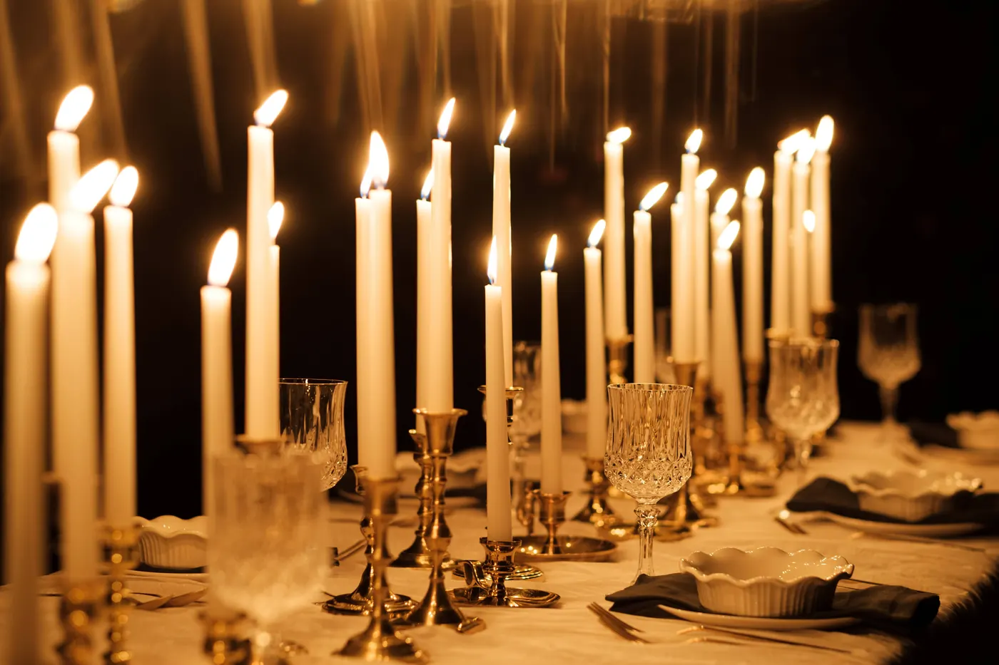 A long candlelit table of tall white tapers in brass candlesticks, with crystal goblets and scalloped bowls on dark napkins