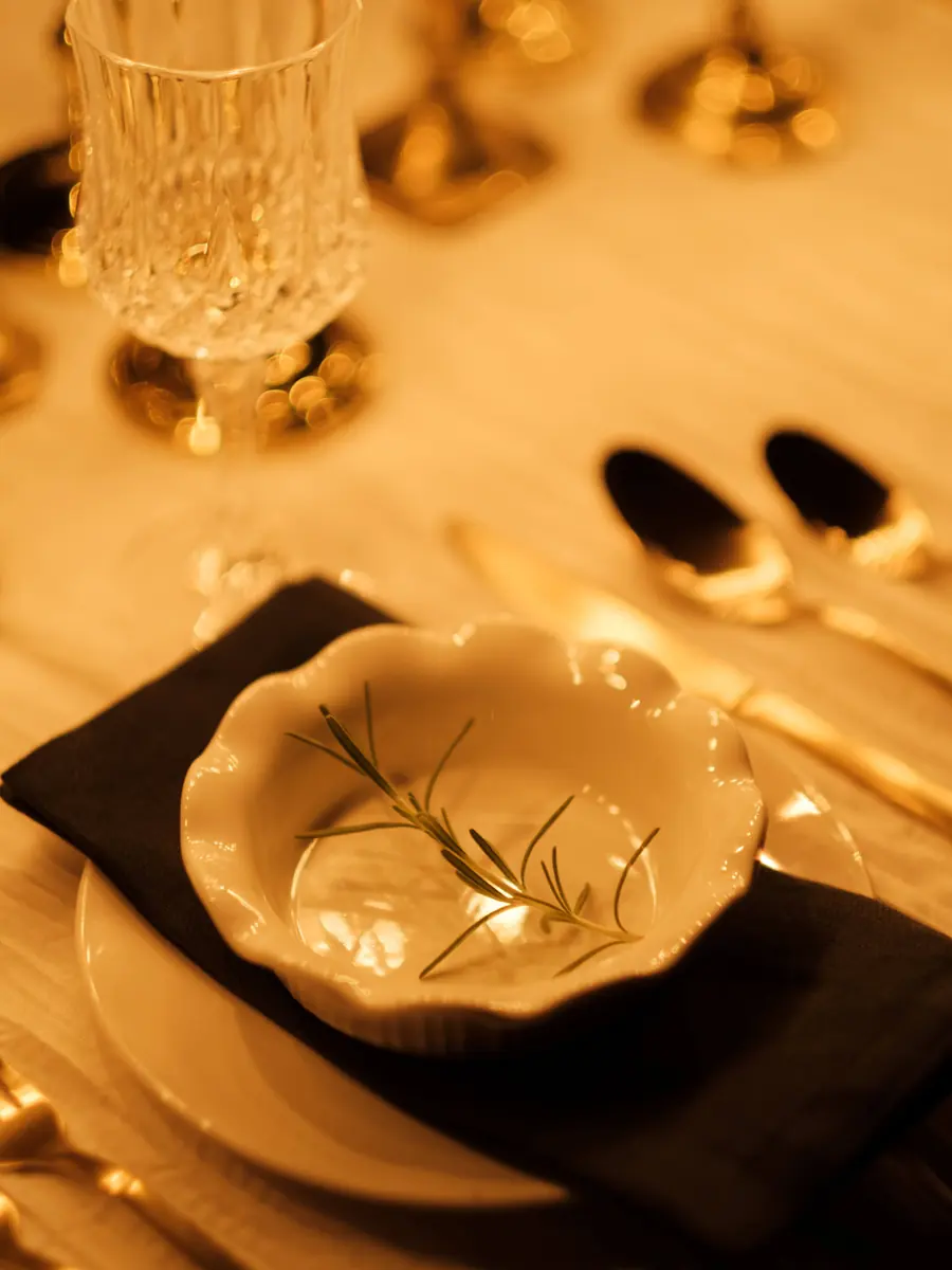 A scalloped bowl with a sprig of rosemary on a dark napkin, with gold flatware