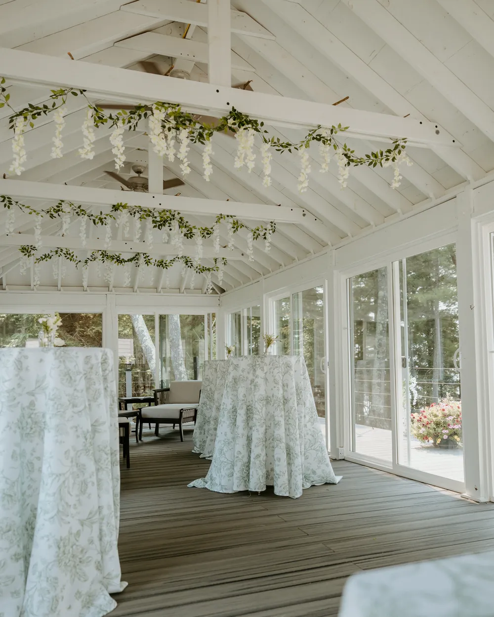 A light-filled sunroom set for a reception, with sage toile tablecloths and greenery garlands